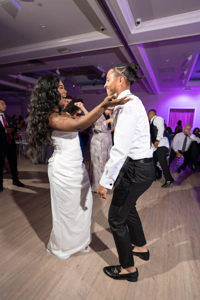 A bride and groom dance together on a wooden floor at a wedding reception, smiling and surrounded by guests in formal attire.
