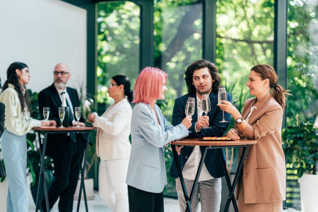 A group of people in business attire stand around cocktail tables, holding drinks and conversing in a bright room with large windows and greenery outside.