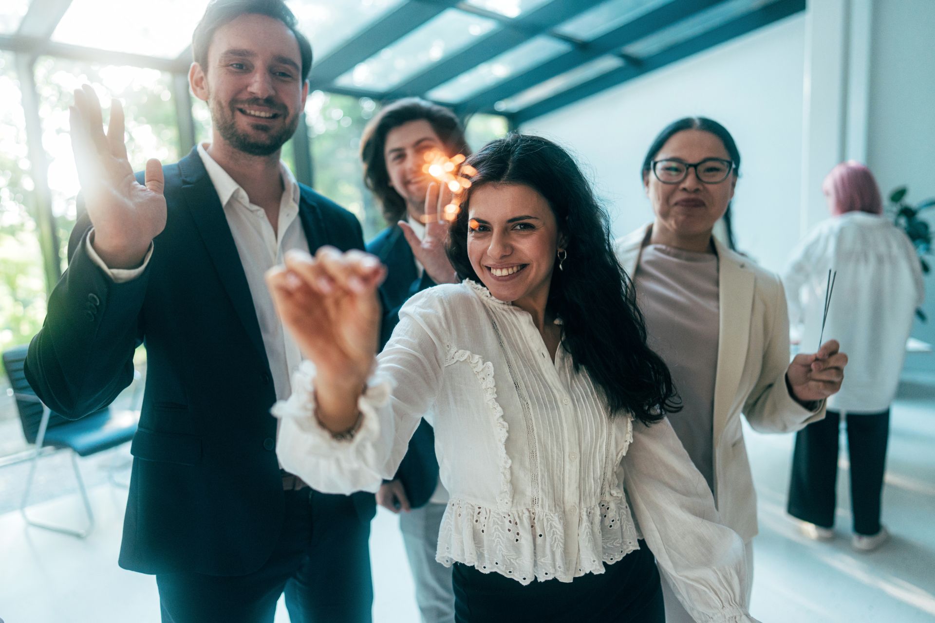Four people indoors, smiling and waving sparklers, with another person in the background; all are dressed in business or smart casual attire.
