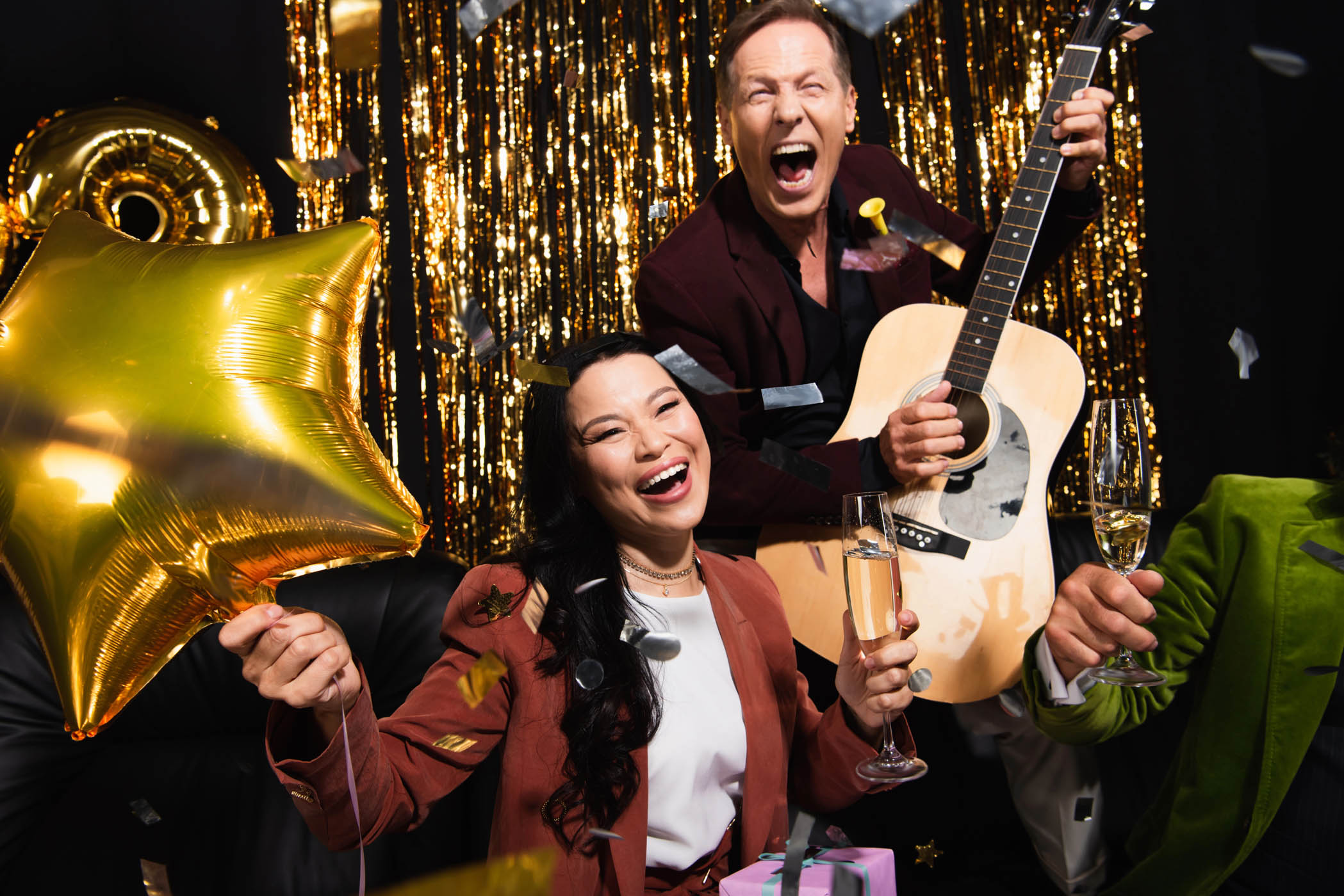 Two people celebrate at a corporate event with gold decorations; one holds a star balloon and champagne, the other plays guitar and laughs as confetti falls.