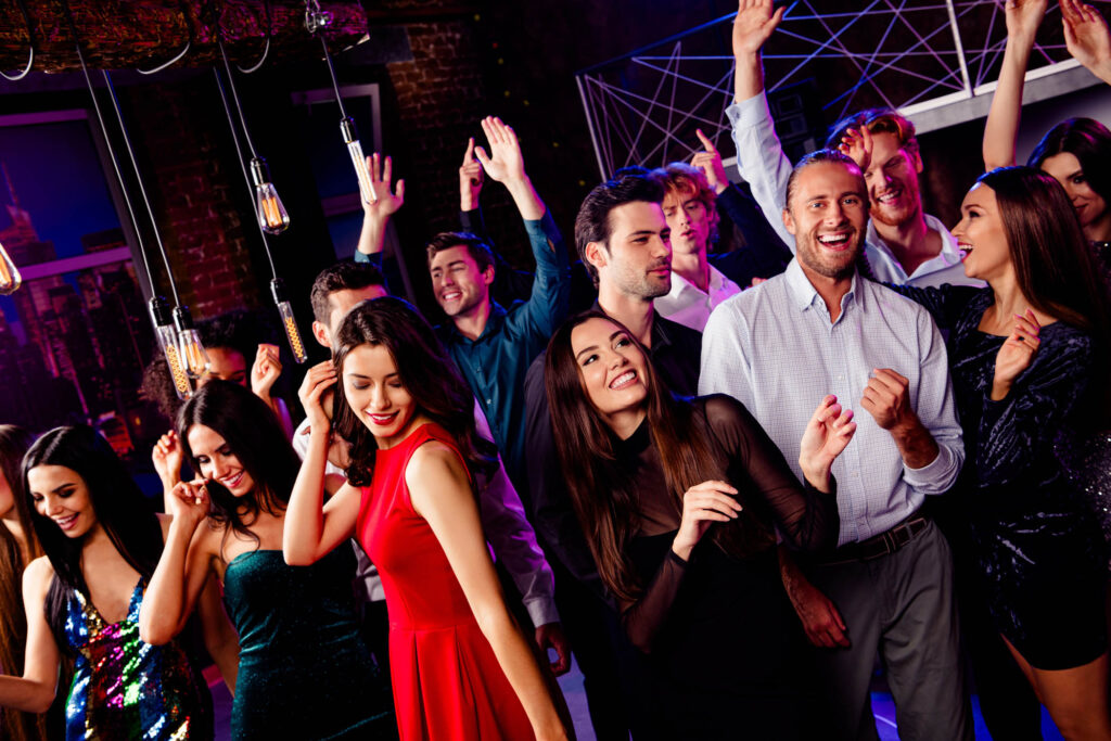 A group of people dressed in party attire are dancing and smiling at an indoor corporate event under hanging lights.
