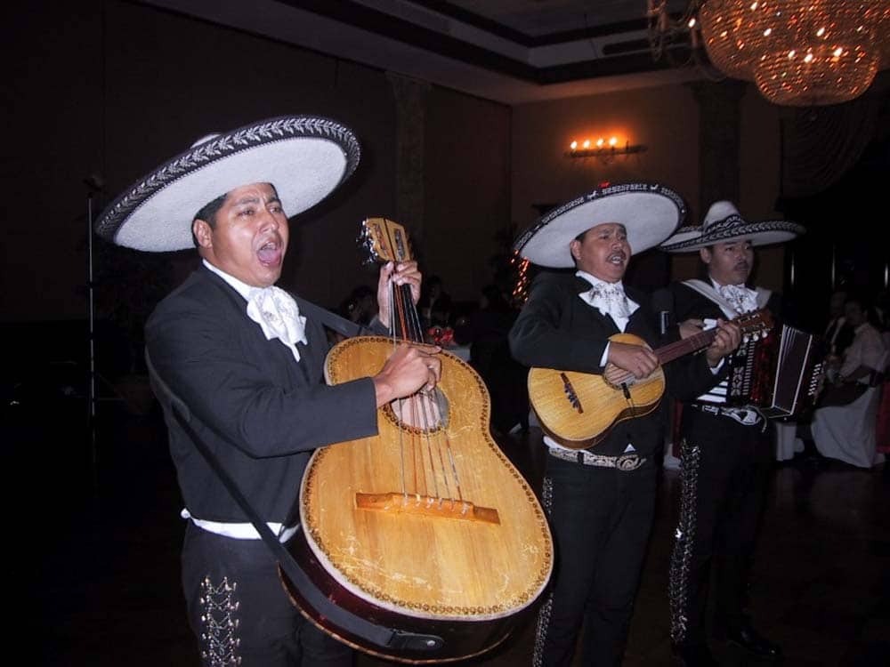 Three mariachi musicians perform indoors, wearing traditional charro suits and sombreros, playing a guitarrón, a vihuela, and an accordion under chandeliers.