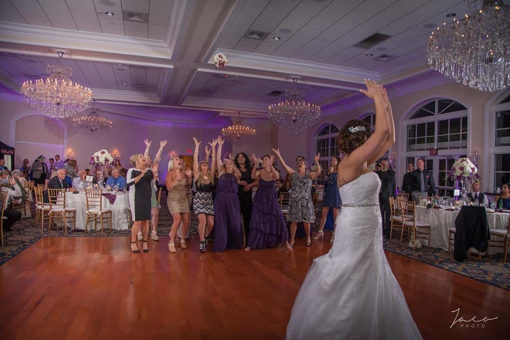 A bride in a white gown tosses her bouquet over her head as a group of women stand ready to catch it at a wedding reception.