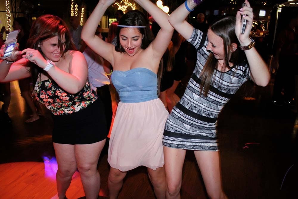 Three young women are dancing together on a dark indoor L.E.D. Dance Floor, smiling and raising their arms, with festive lights in the background.