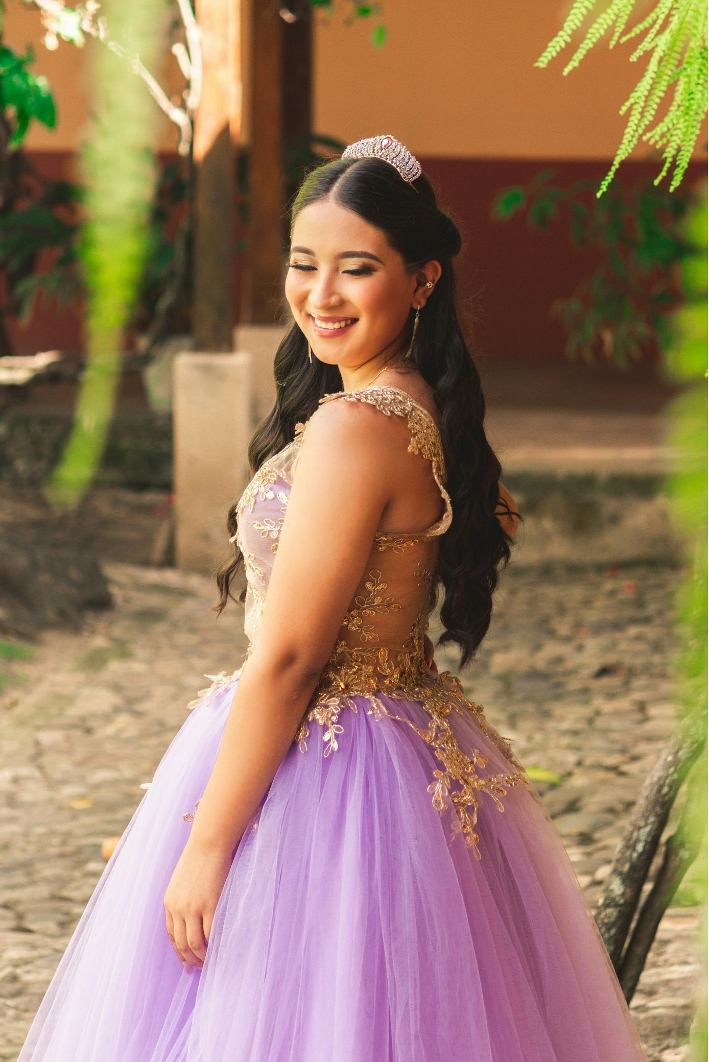 Young woman in a lavender and gold formal dress stands outdoors on a cobblestone path, smiling and looking over her shoulder.