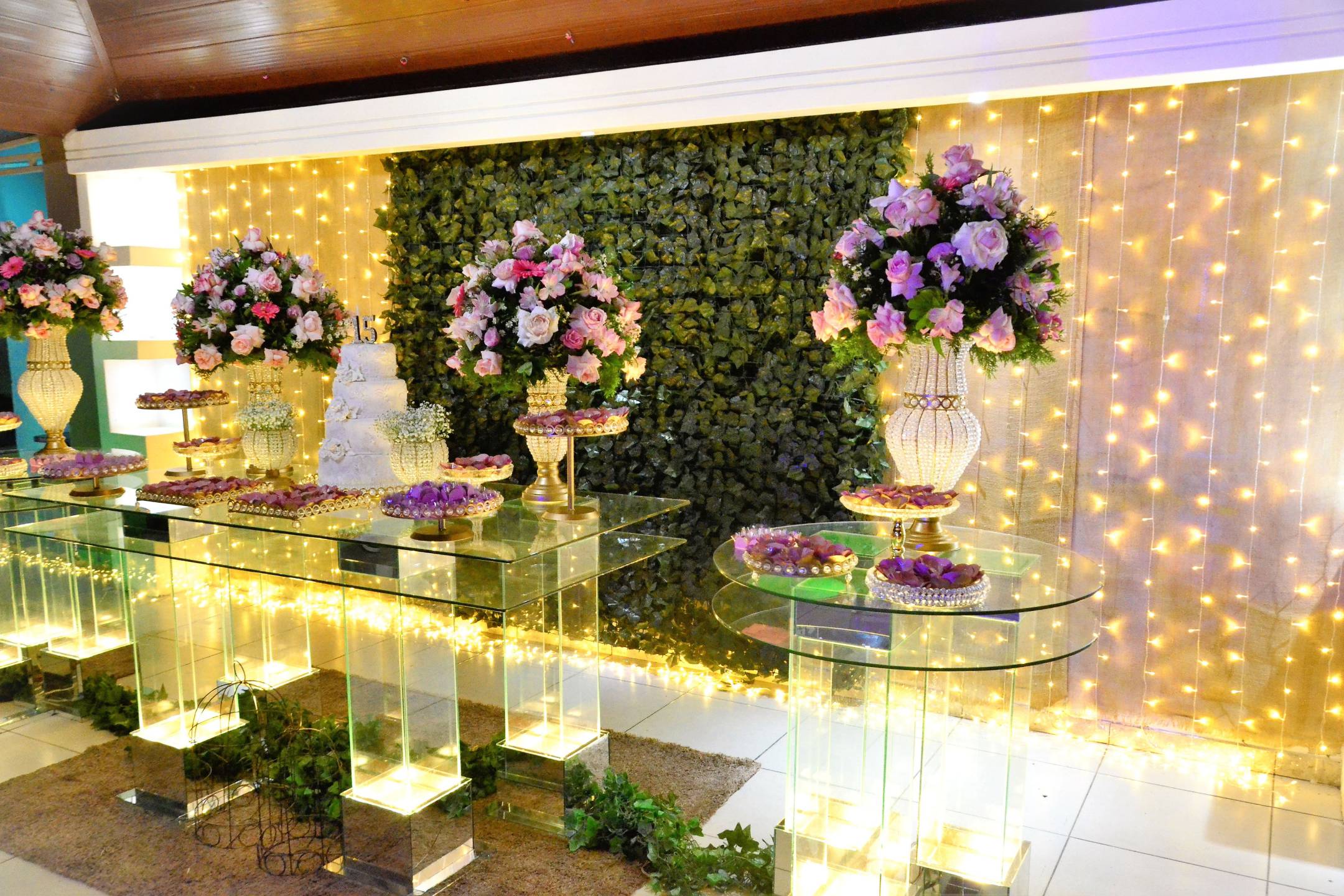 A decorated event table with glass surfaces, floral arrangements, a tiered cake, assorted desserts, and a backdrop of greenery and string lights.