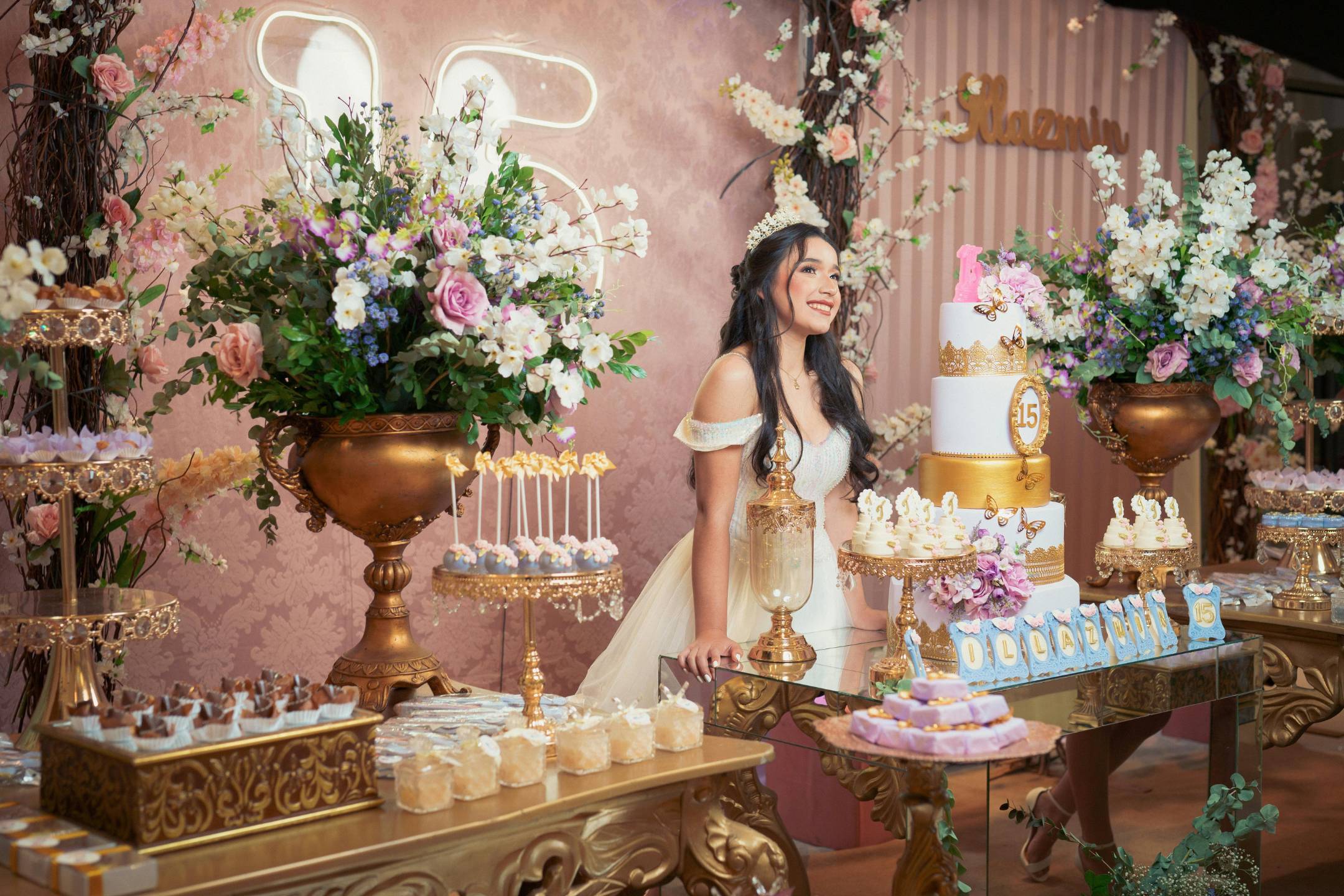 A young woman in a formal dress stands smiling at a lavishly decorated table with a large cake, desserts, and floral arrangements at a quinceañera celebration.