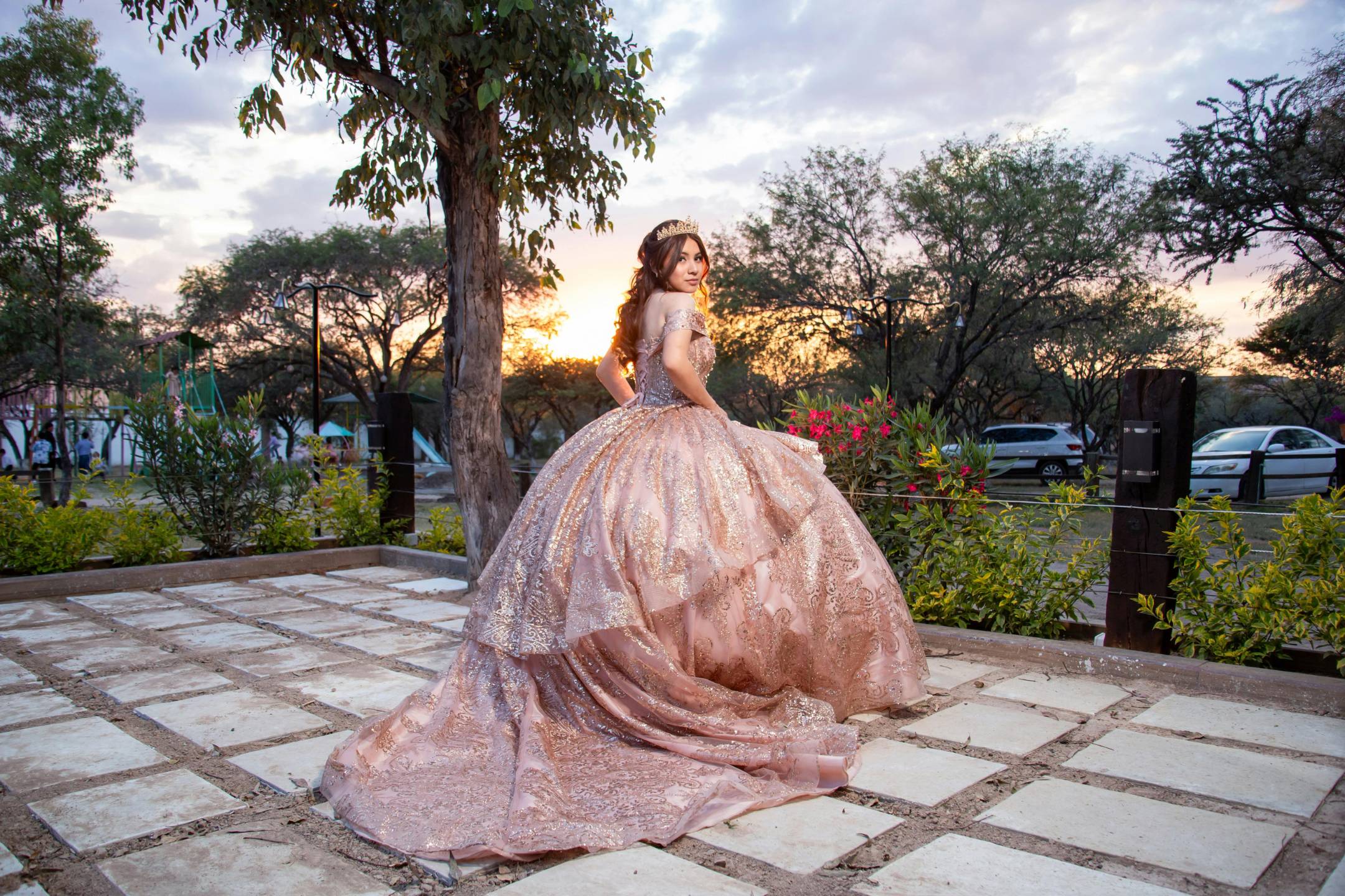A young woman in a jeweled ball gown stands outdoors on a tiled patio at sunset, looking over her shoulder toward the camera.