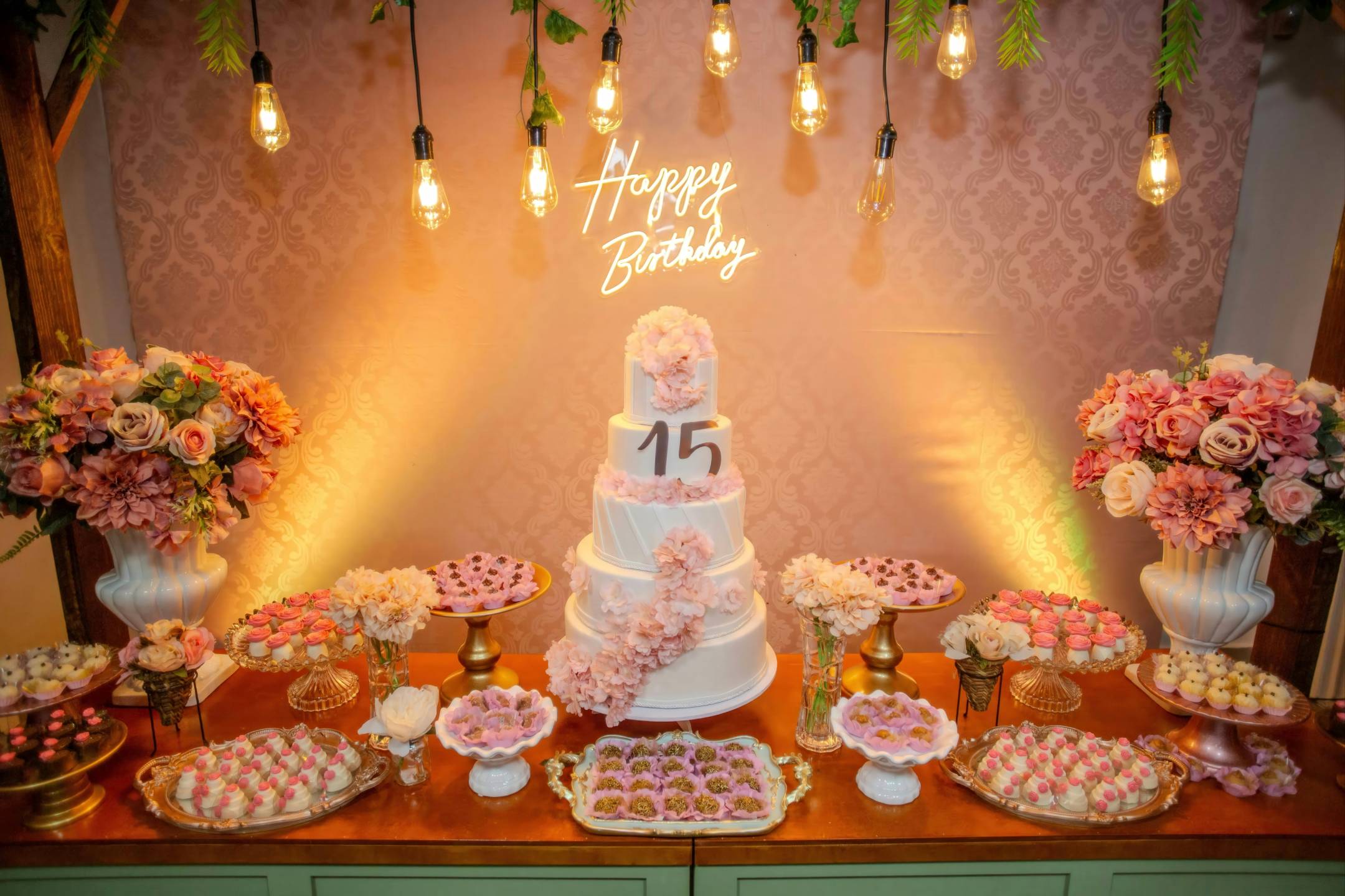 A decorated birthday dessert table with a white cake topped with “15,” surrounded by flowers, assorted sweets, and a “Happy Birthday” neon sign in the background.