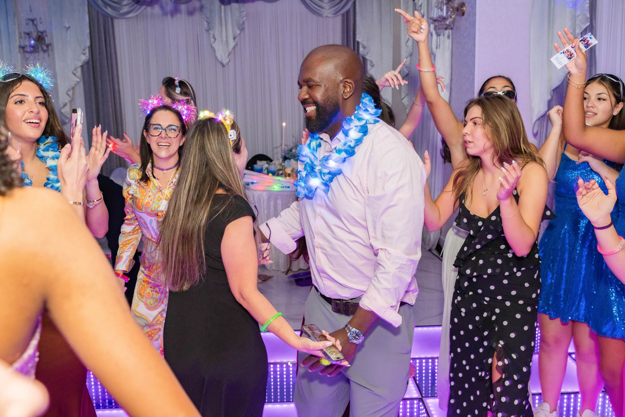A group of people in festive attire dance and celebrate together on a lit-up floor at an indoor party or event.