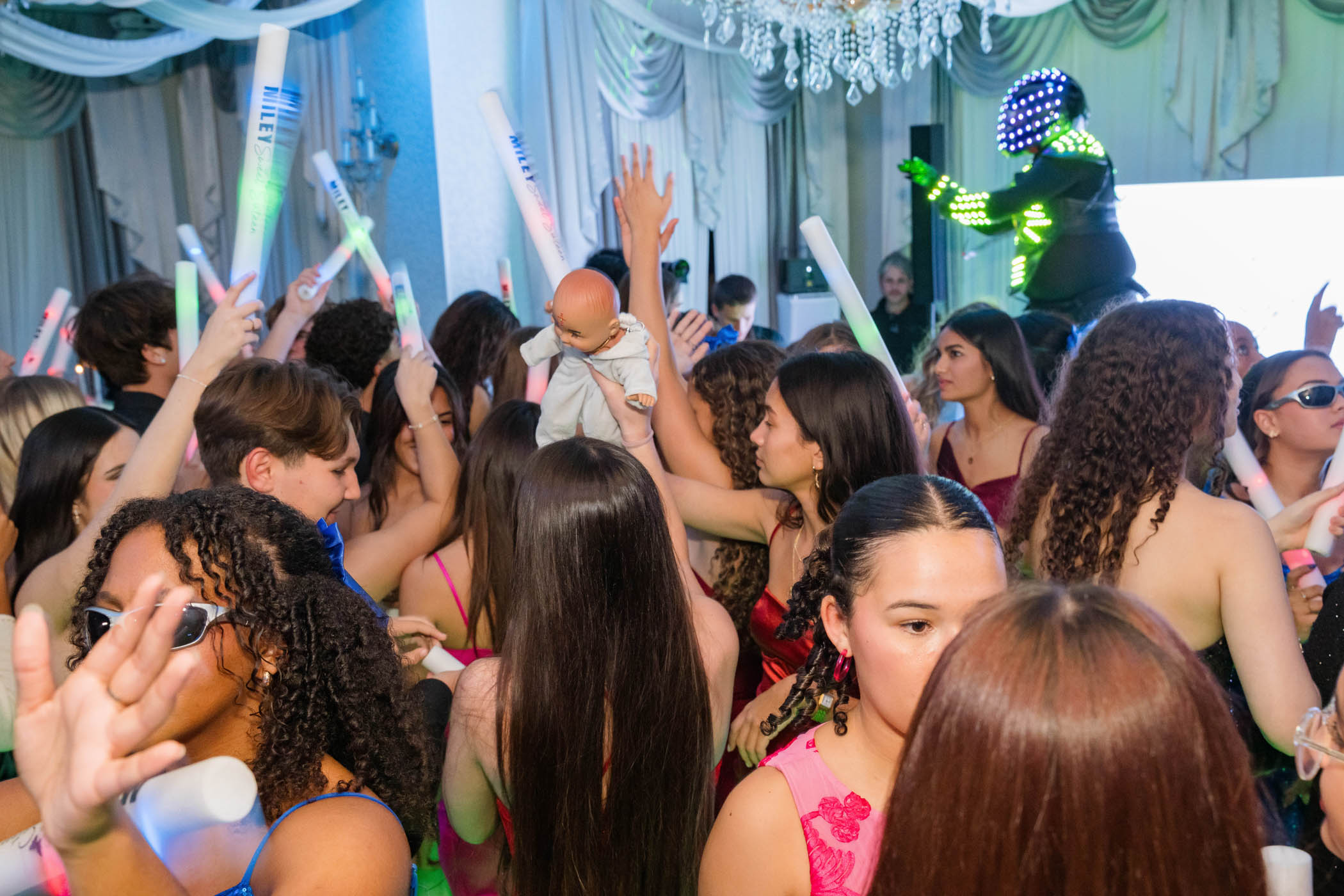 A group of people dance at a crowded indoor party; some hold light sticks and one person raises a baby doll while a performer in LED costume stands in the background.