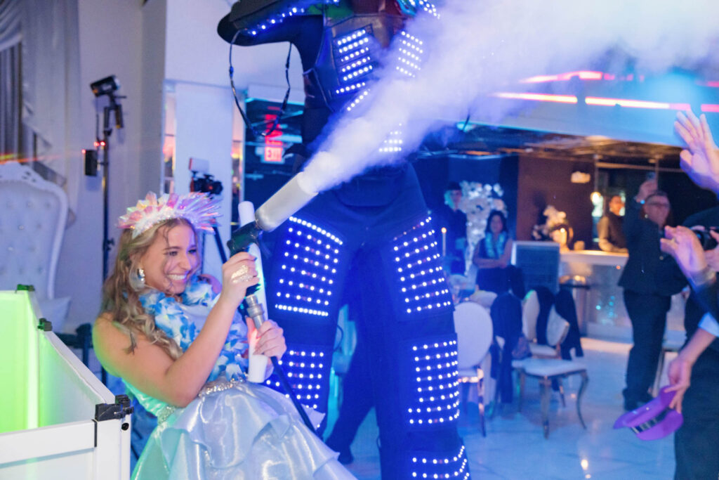 A woman in a blue dress and flower crown smiles while spraying fog from a machine next to a person in a glowing LED suit at a party with people in the background.