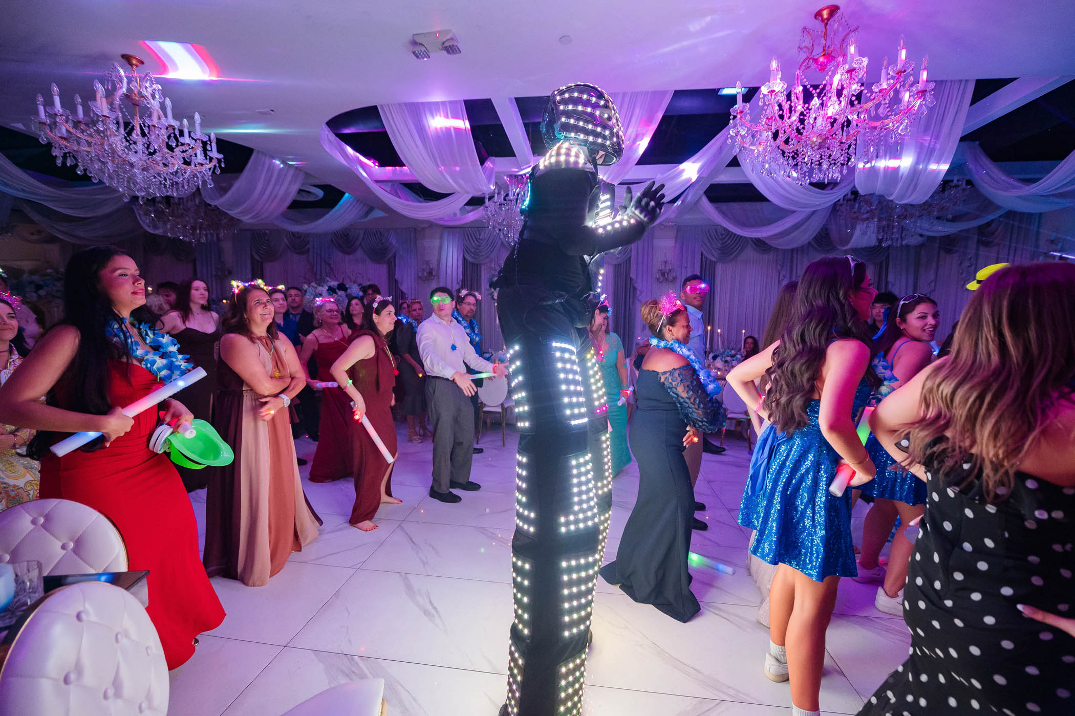 A performer in an illuminated LED suit entertains a crowd of people dressed in formal and festive attire at an indoor event with chandeliers and draped decorations.