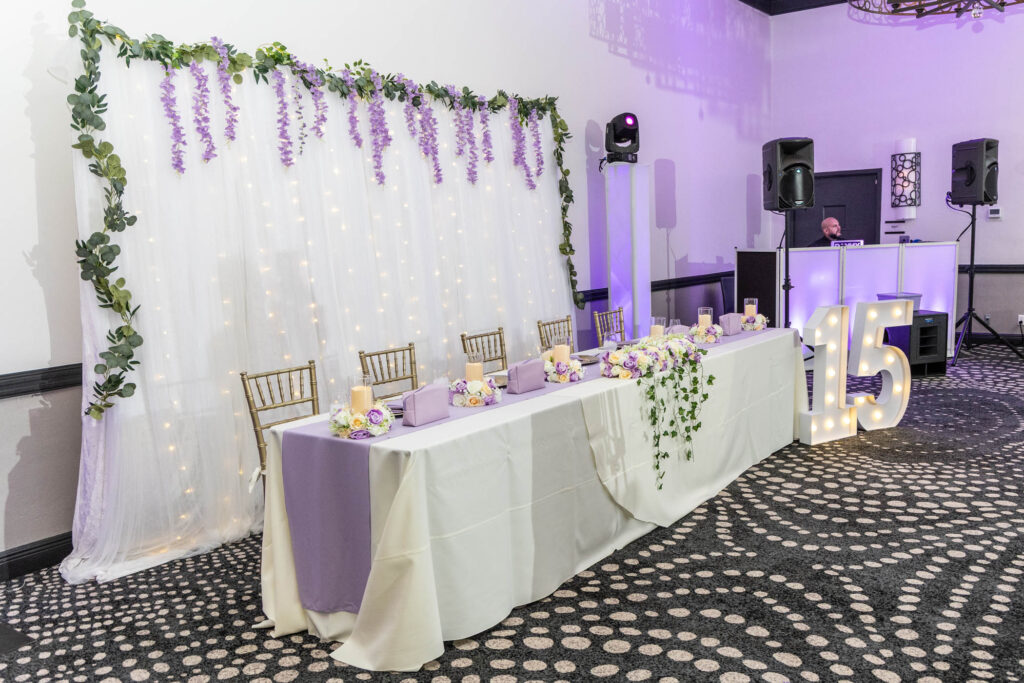 A long, white and lavender decorated table sits in front of a backdrop with hanging flowers and fairy lights; large illuminated "15" numbers are beside a DJ booth in the corner.