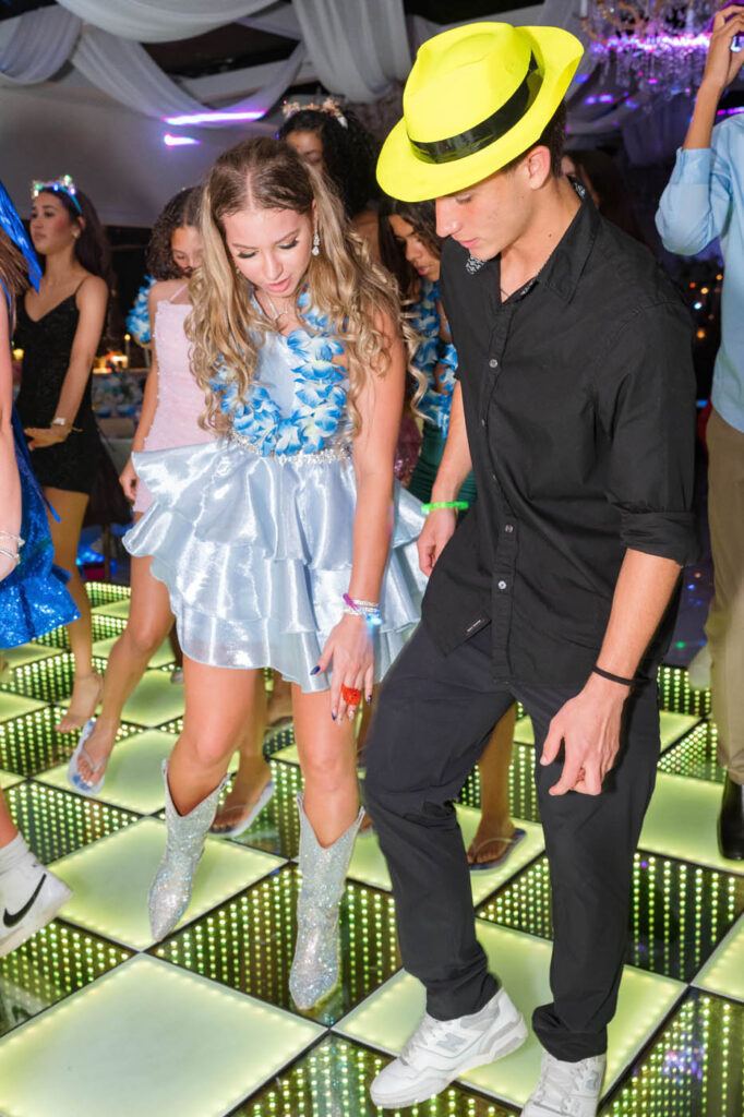 Two people dance on an illuminated floor at a party; the woman wears a blue dress and boots, the man wears black clothes and a yellow hat. Other guests are visible in the background.