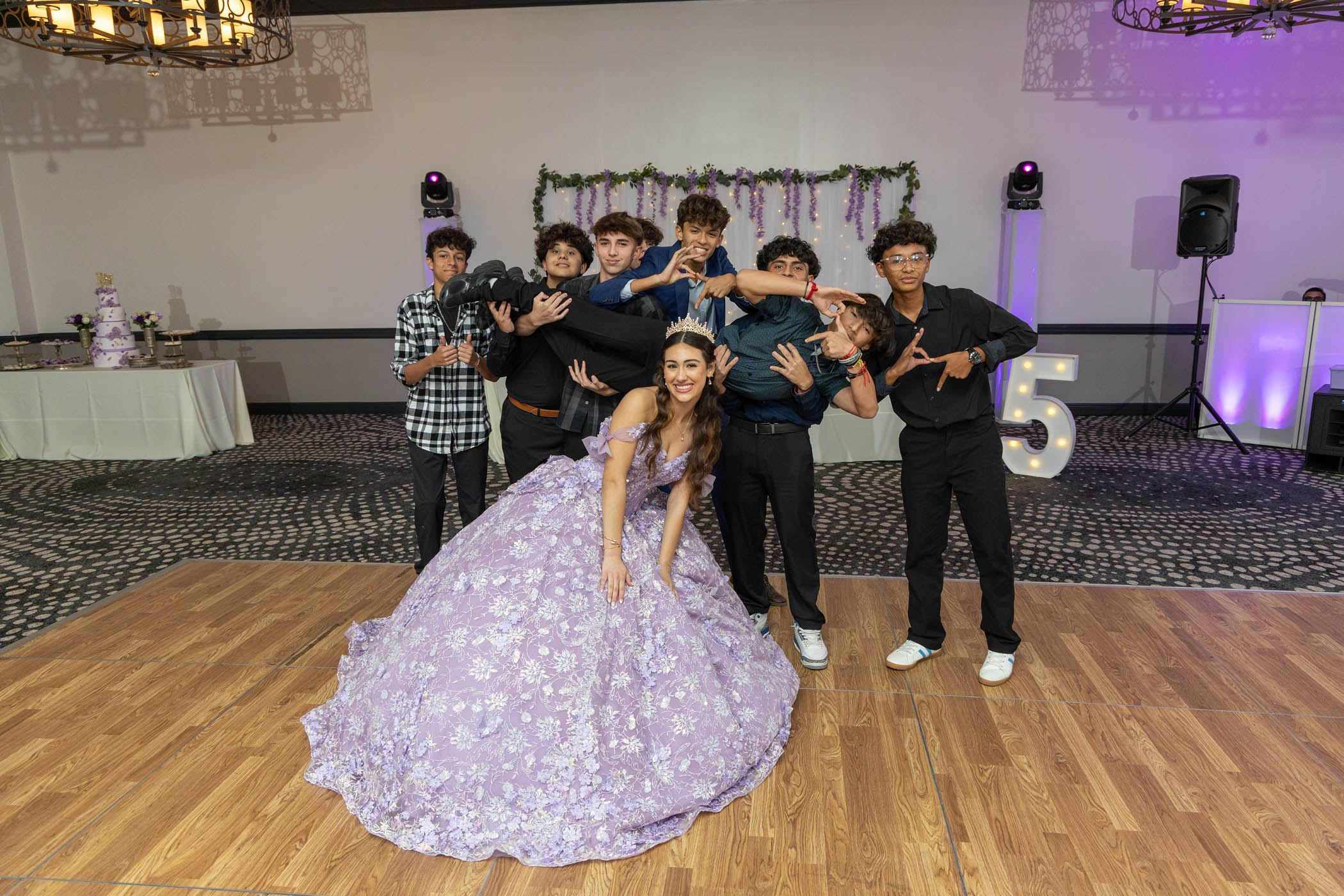 A young woman in a lavender ball gown poses in front of a group of boys, one of whom is being held horizontally by others, at an indoor event with floral decorations and a large light-up "15.