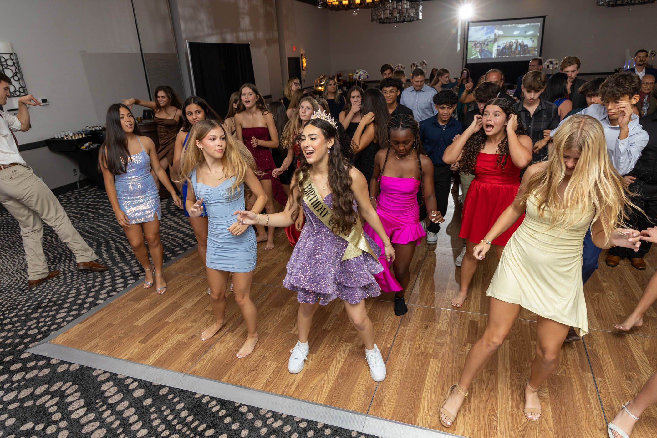 A group of young people dance together on a wooden floor at an indoor party or event, with some wearing formal dresses and a girl in a sash leading the group.