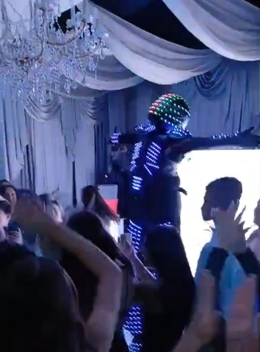 Performer in an LED light suit stands with arms outstretched on a stage, surrounded by people dancing under draped fabrics and chandeliers.