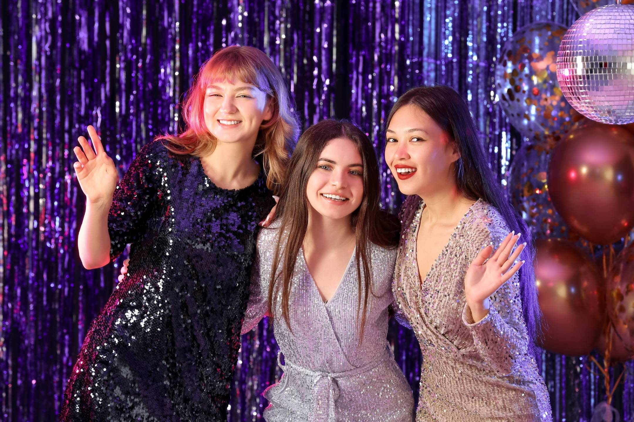 Three women in sparkly dresses smile and pose together at a party, with metallic streamers, balloons, and a  ball in the background.