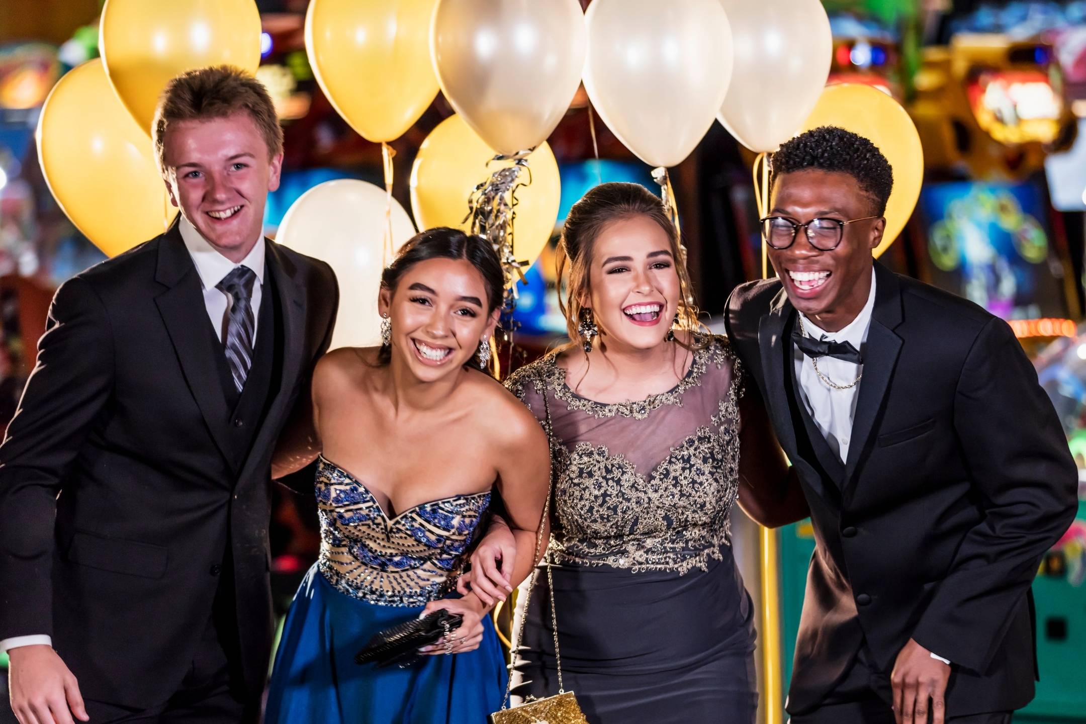 Four young adults dressed formally stand close together, smiling and laughing, with gold and white balloons in the background.