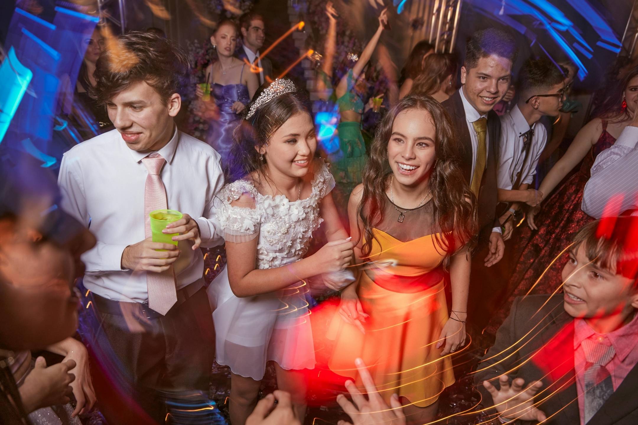 A group of young people dressed for a formal event dance and smile among colorful lights at a party.