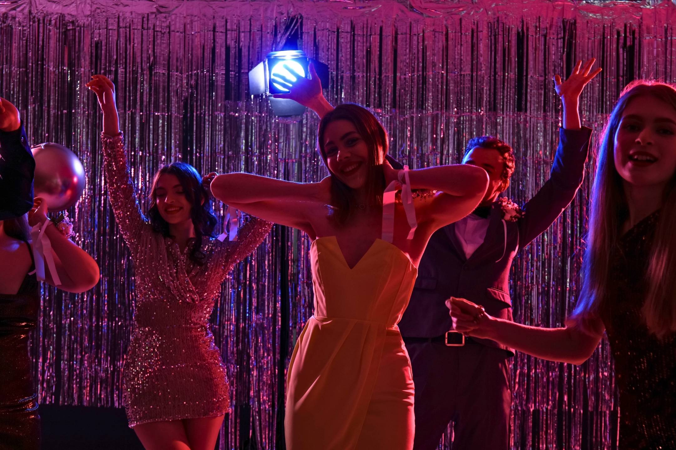 A group of young people dressed formally dance and pose under pink lighting in front of a metallic tinsel backdrop at a party or prom.