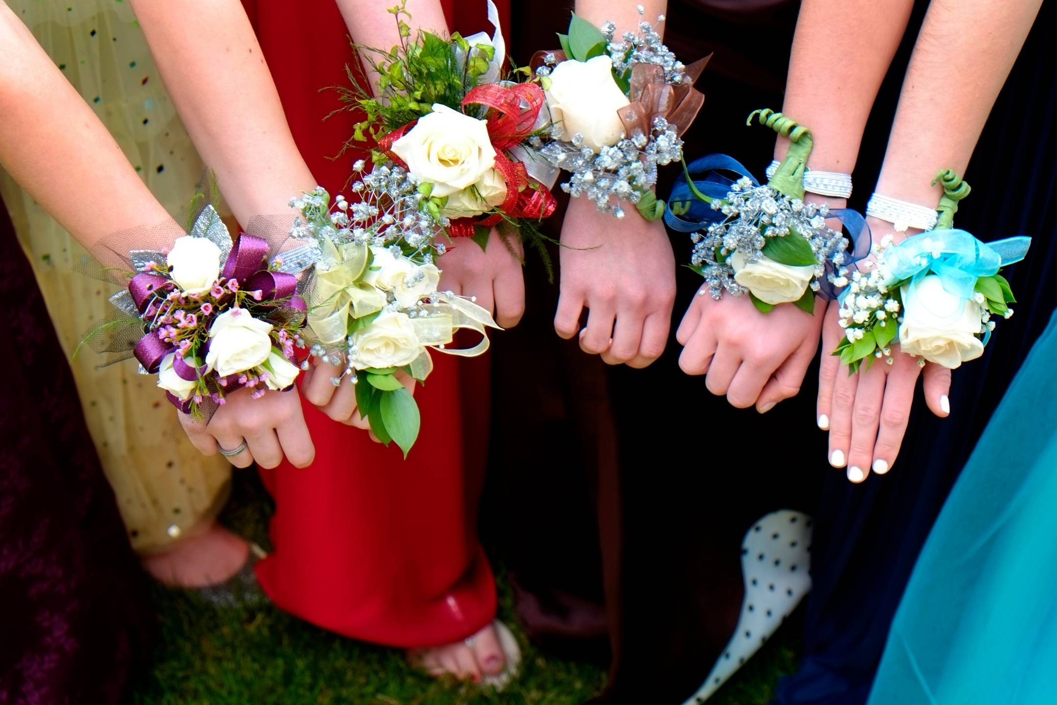 Four people wearing formal dresses show corsages with white flowers and colored ribbons on their wrists, standing close together outdoors.