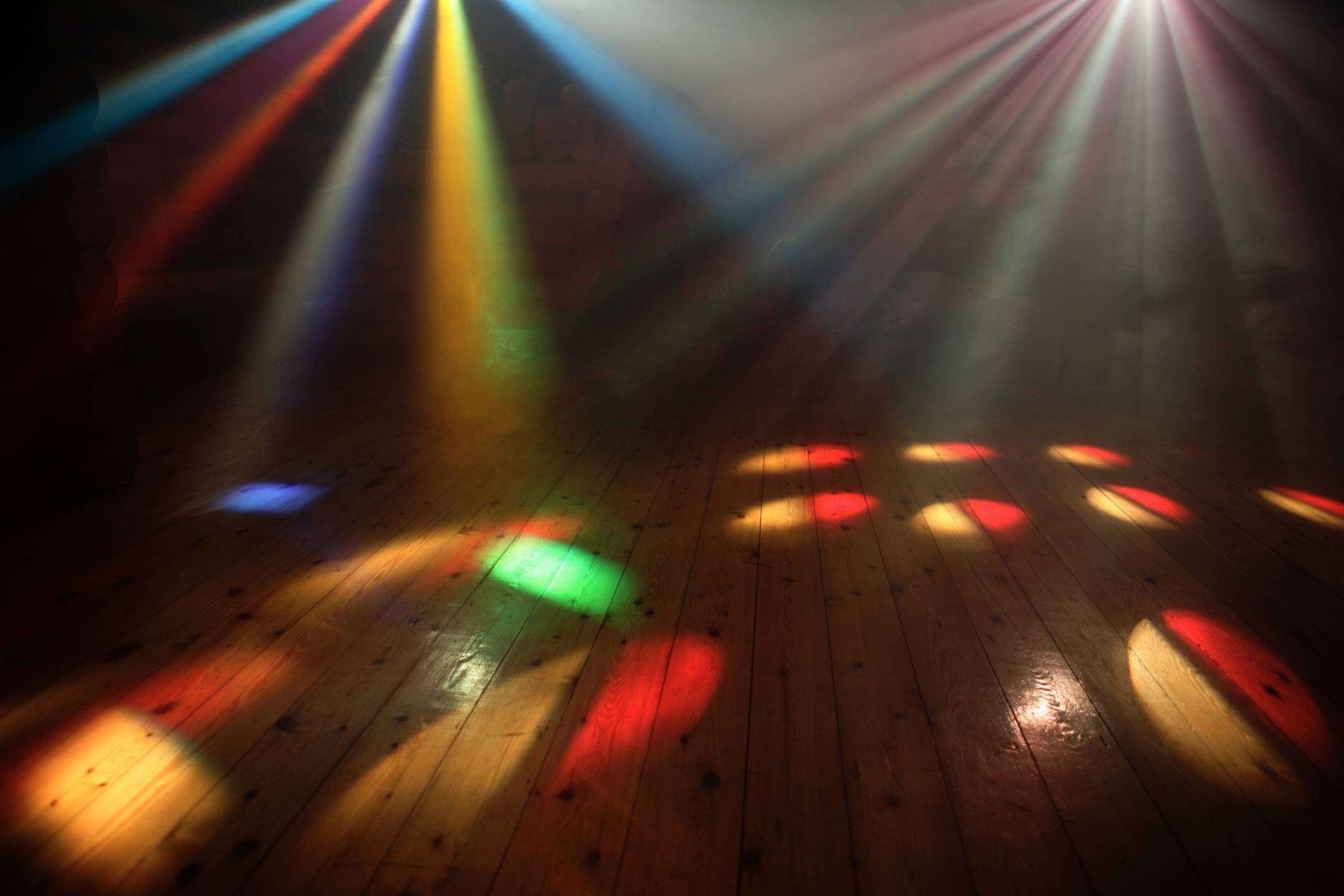 Colorful stage lights cast beams and circular patterns onto a wooden floor in an empty, smoky room.