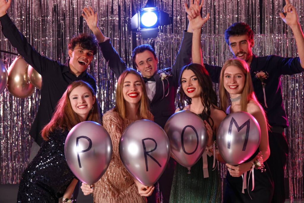 Six young people at a prom celebrate in front of a silver tinsel backdrop, holding balloons spelling "PROM" and smiling at the camera.
