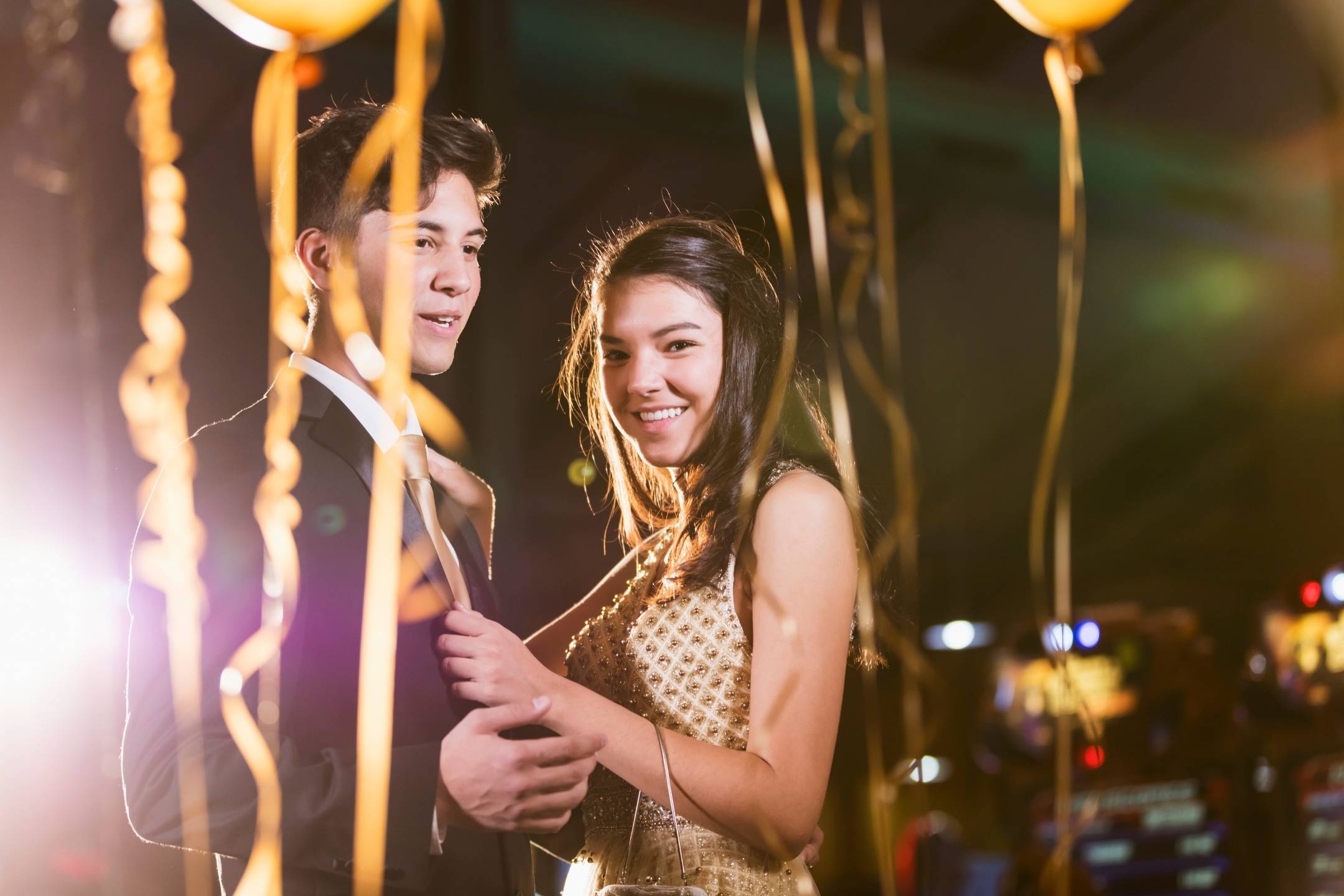 A young couple in formal attire stands together at a decorated event, surrounded by golden balloons and streamers, smiling at the camera.