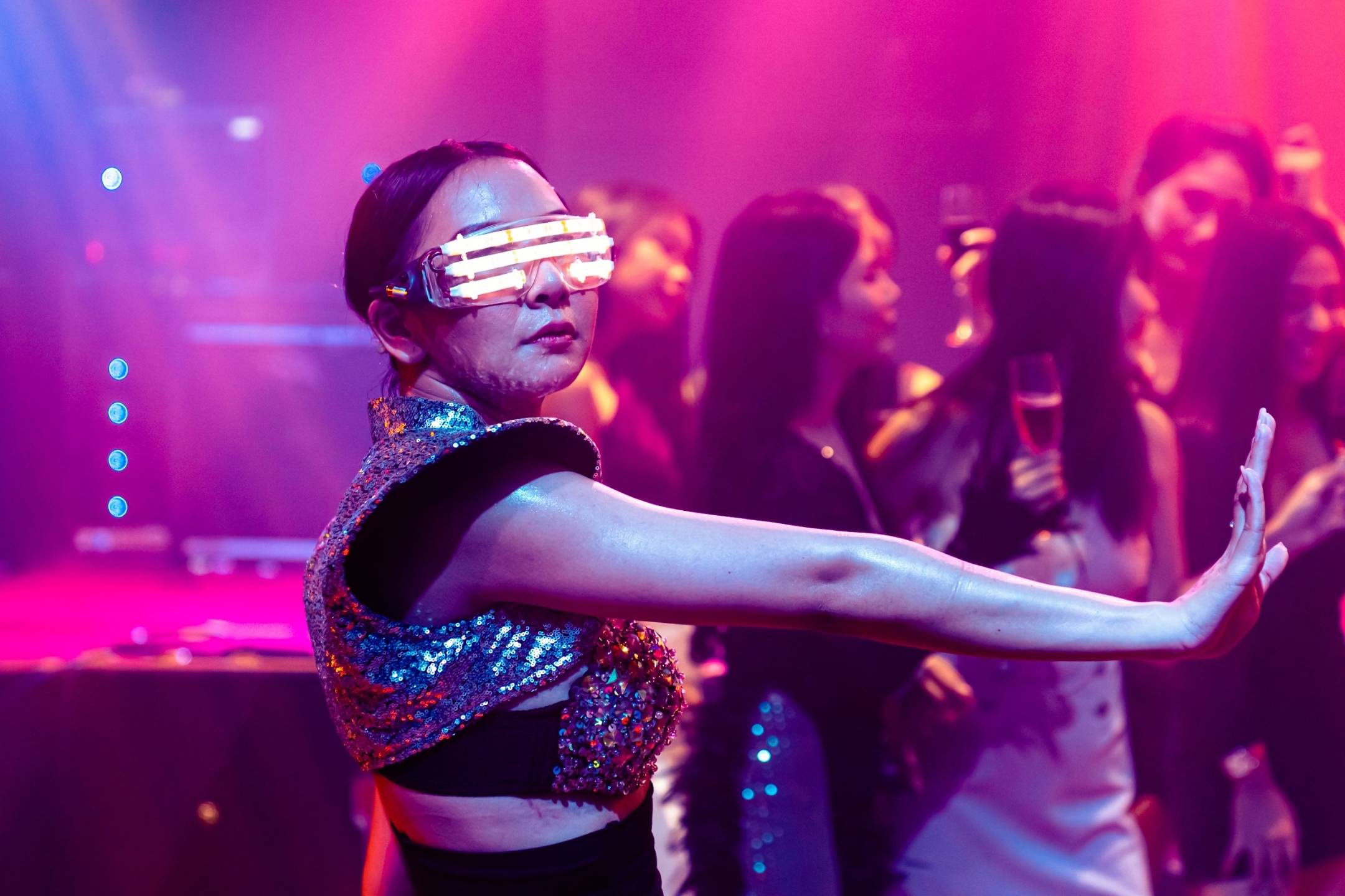 A person wearing futuristic LED glasses and a sequined outfit poses with an outstretched arm at a party with people in the background under colorful lights.