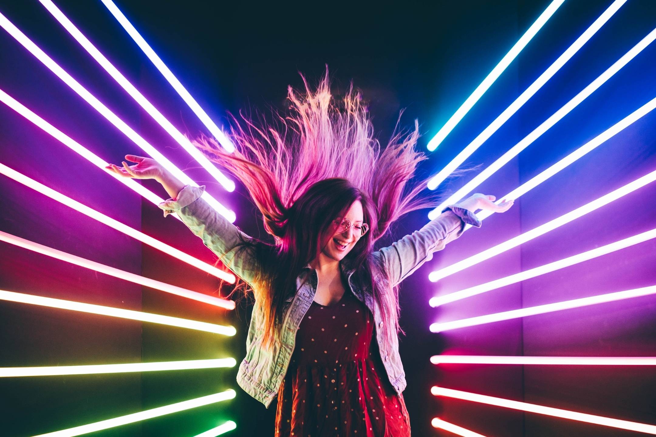 A person with long hair wearing glasses and a dress poses with arms raised among colorful neon light beams in a dark room.