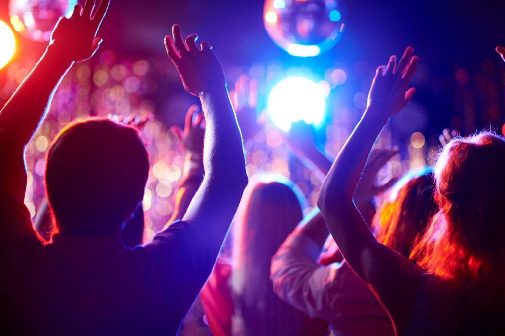 A group of people with raised hands dance under colorful lights at a crowded party or nightclub.