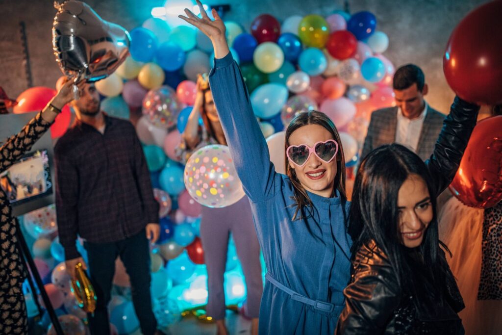 People at a party, surrounded by colorful balloons; one woman in heart-shaped sunglasses and a blue outfit raises her arm and smiles at the camera.