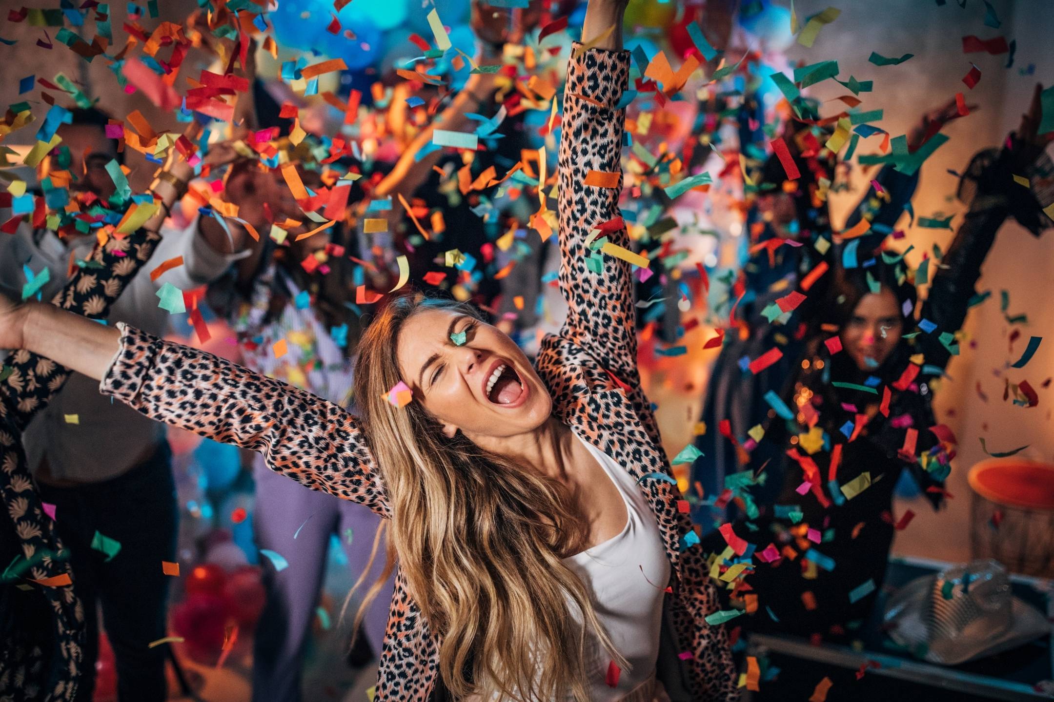 A woman in a leopard print jacket celebrates with arms raised, surrounded by colorful confetti and people at a lively party.