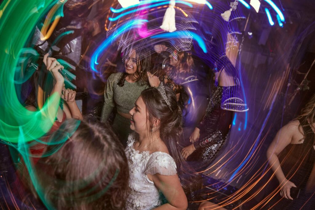 Several people dance at a party under colorful, swirling lights, with one person in a white dress in the center of the frame.