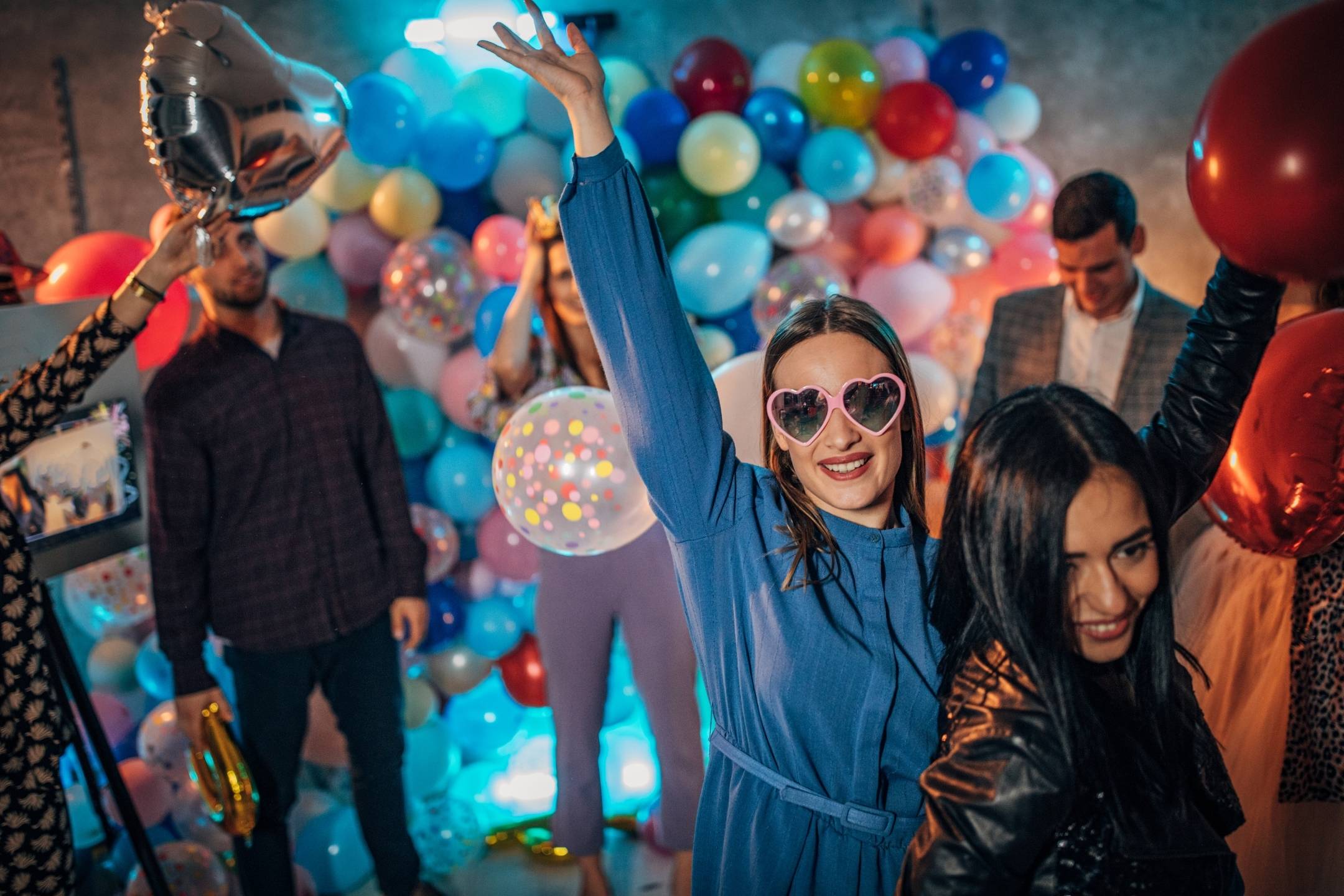 Group of people at a party with colorful balloons in the background; a woman in heart-shaped sunglasses raises her arm and smiles at the camera.