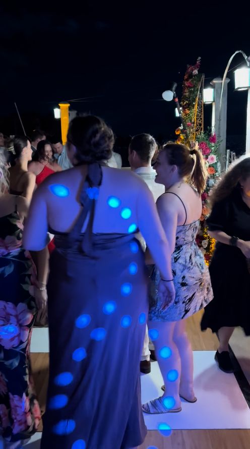 A group of people stand on a L.E.D. Dance Floor at night, illuminated by blue lights, with floral decorations and lamps in the background.
