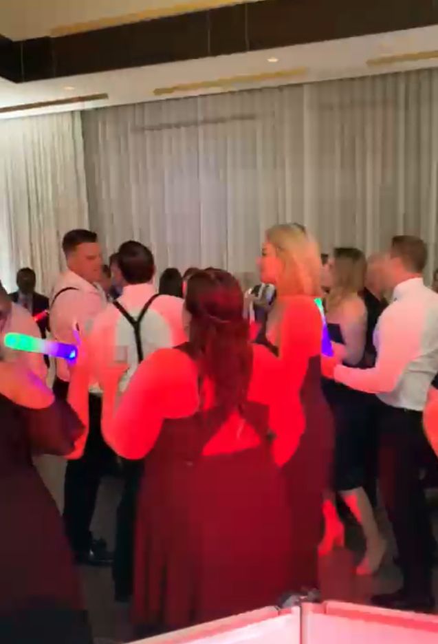 A group of people in formal attire dance indoors, some holding glowing light sticks, with red lighting illuminating the scene.