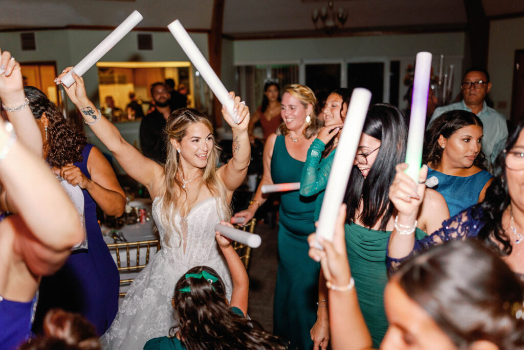 A bride in a white dress celebrates with guests, capturing the excitement people learn about when experiencing a lively indoor wedding reception with glowing foam sticks and dancing.