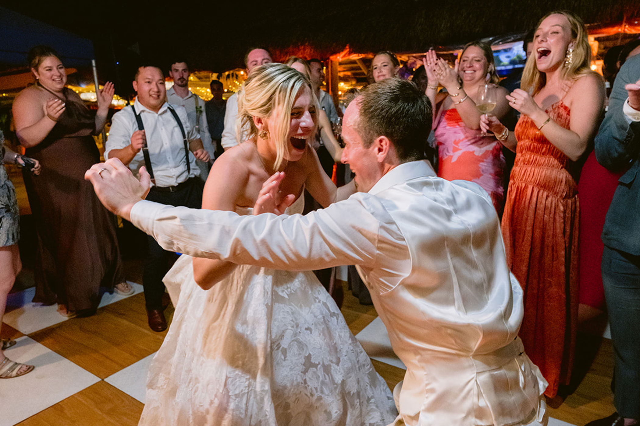 A bride and groom dance energetically on a checkered floor, surrounded by cheering guests at a lively wedding reception, brought to life by top-tier wedding DJ Services.