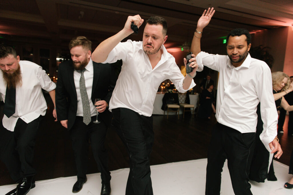 Four men in dress shirts and ties dance energetically at an indoor event, holding drinks and raising their arms, with tables and guests visible in the background.