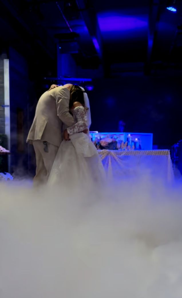 A bride and groom in formal attire embrace on a smoke-filled L.E.D. Dance Floor, with a table and blue lighting in the background—creating the perfect moment with professional wedding DJ services.