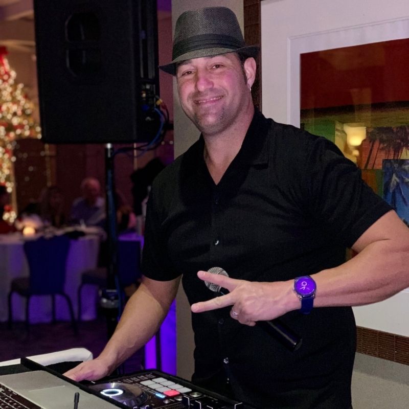 A man wearing a black shirt and hat holds a microphone and makes a peace sign while standing at a DJ booth during an indoor event.