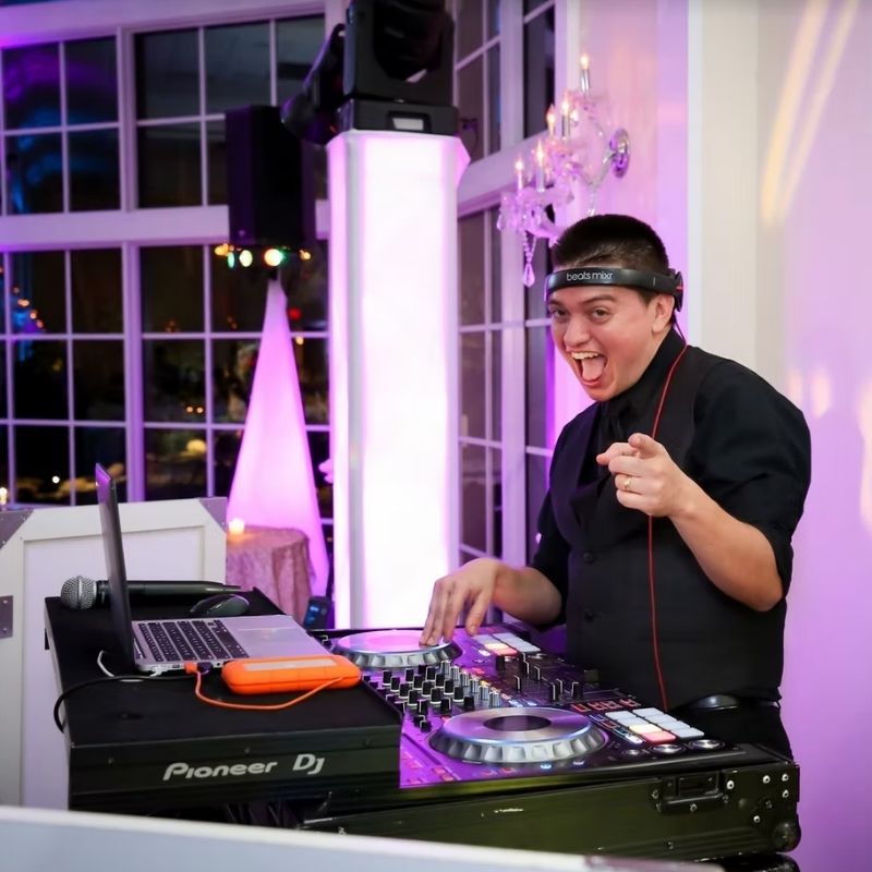 A DJ wearing headphones and a black vest stands behind a mixing console, smiling and pointing towards the camera in a brightly lit room.