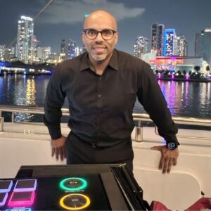 A man in a black shirt stands on a boat at night with a city skyline and colorful lights in the background. A DJ controller is visible in the foreground.