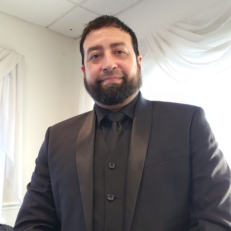 A man with a beard and short dark hair wearing a black suit, shirt, and tie stands indoors with white curtains and ceiling tiles in the background.