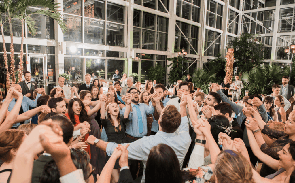 A large group of people hold hands in a circle and sing or shout together at an indoor event with glass walls and tropical plants.