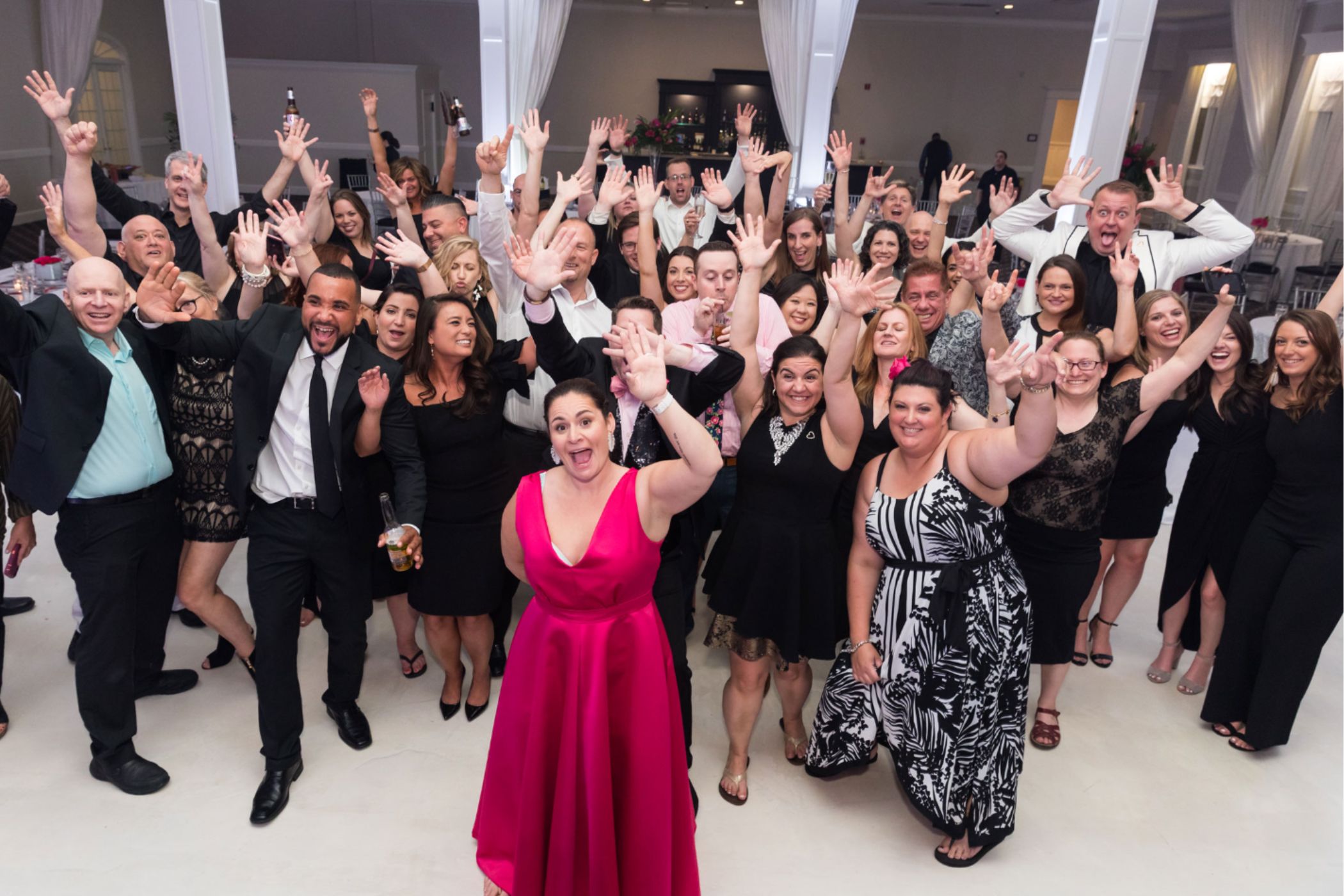 A large group of people dressed in formal attire pose together indoors, smiling and raising their hands toward the camera, enjoying the lively atmosphere provided by Wedding DJ Services.