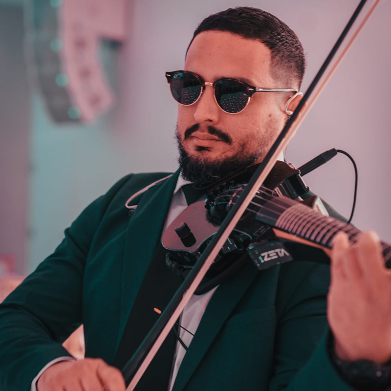 A man wearing sunglasses and a suit is playing an electric violin indoors.
